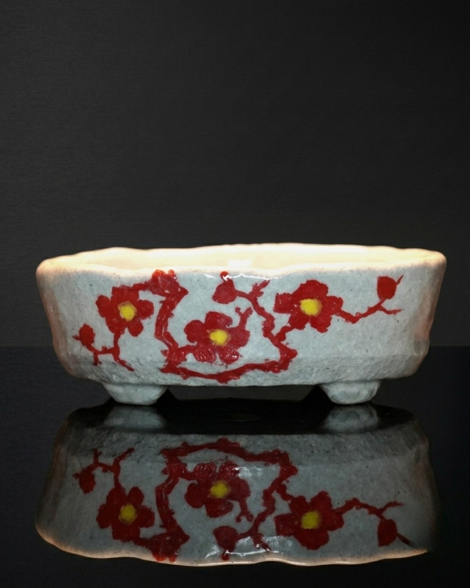 An Echizen Bunzan bonsai pot: red plum blossoms and branches painted over a white glaze, photographed on black with a reflection